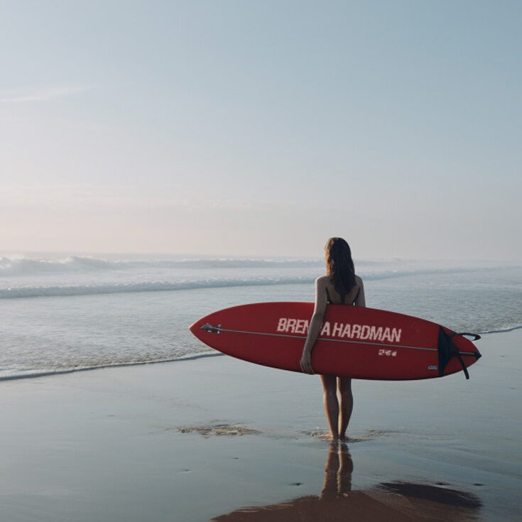 Photography of a surfer holding a red board, illustrating Brenna Hardman's distinct passions such as financial advising and spotting whales sea.